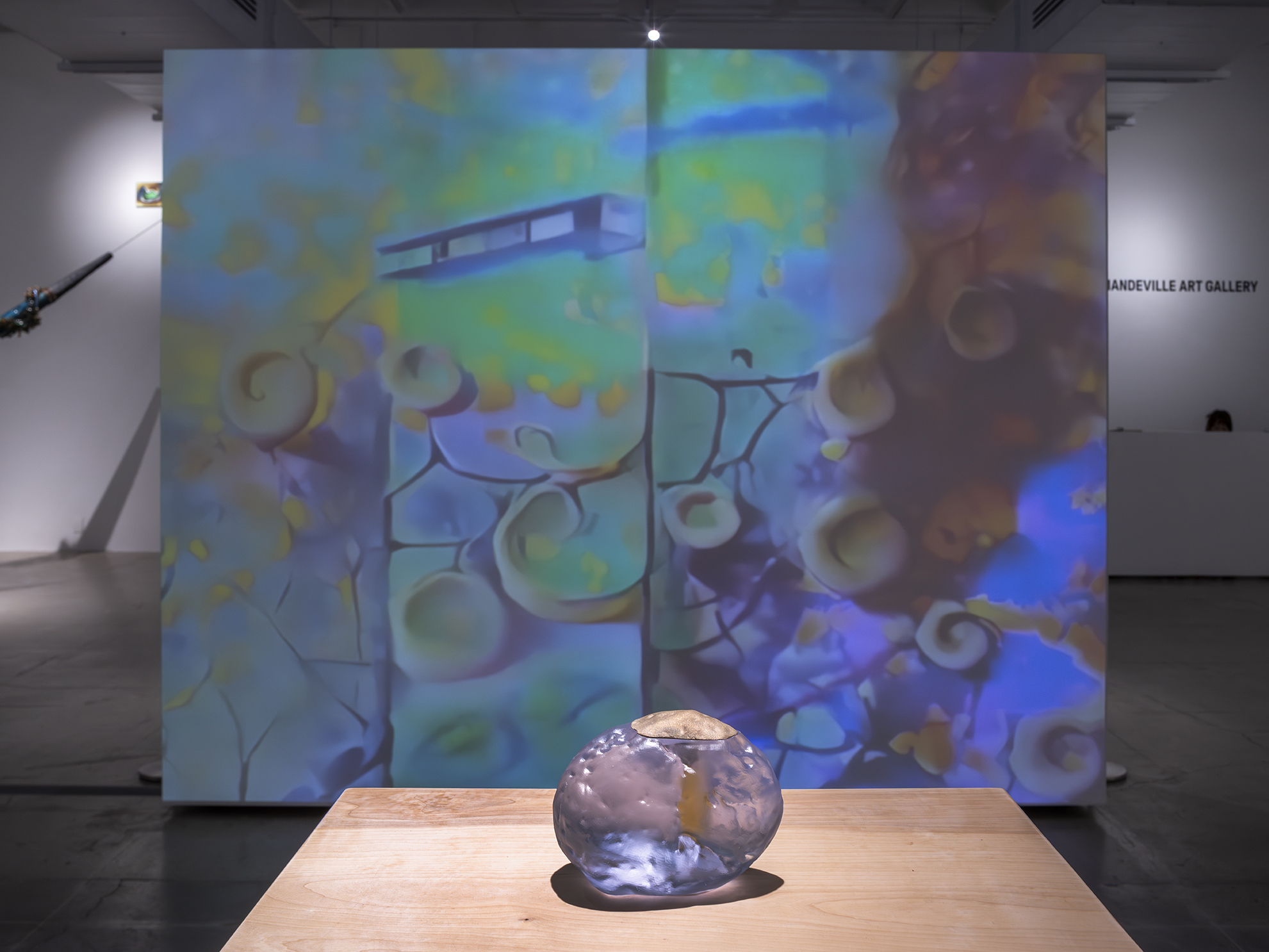 A vibrant, abstract animation with swirling patterns and soft colors is projected in an art gallery. A translucent glass sculpture sits on a wooden table.