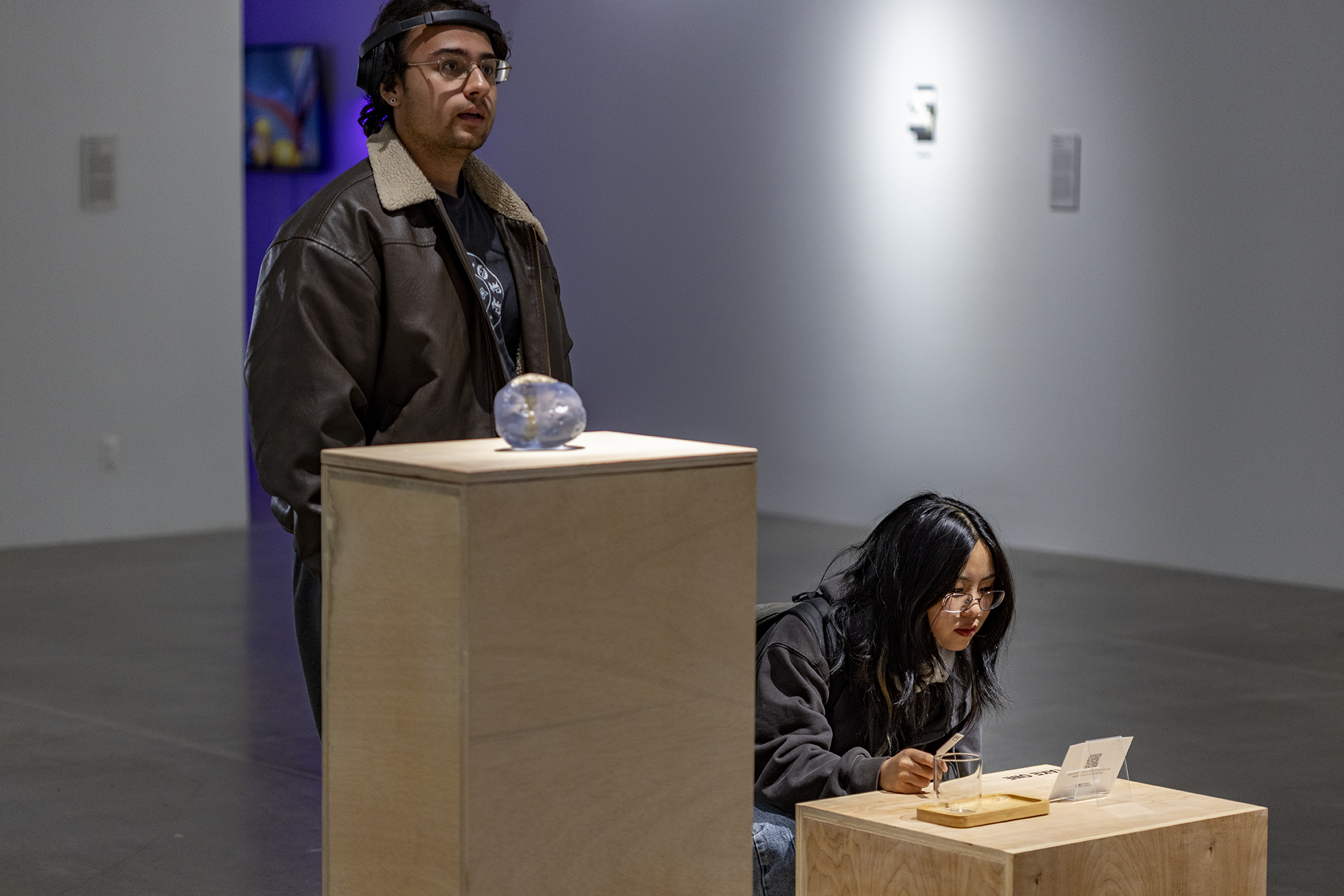 A man in a leather jacket observes a glass orb on a wooden pedestal, while a woman kneels nearby, focused on a small wooden cube with a note. The gallery setting is serene and contemplative.